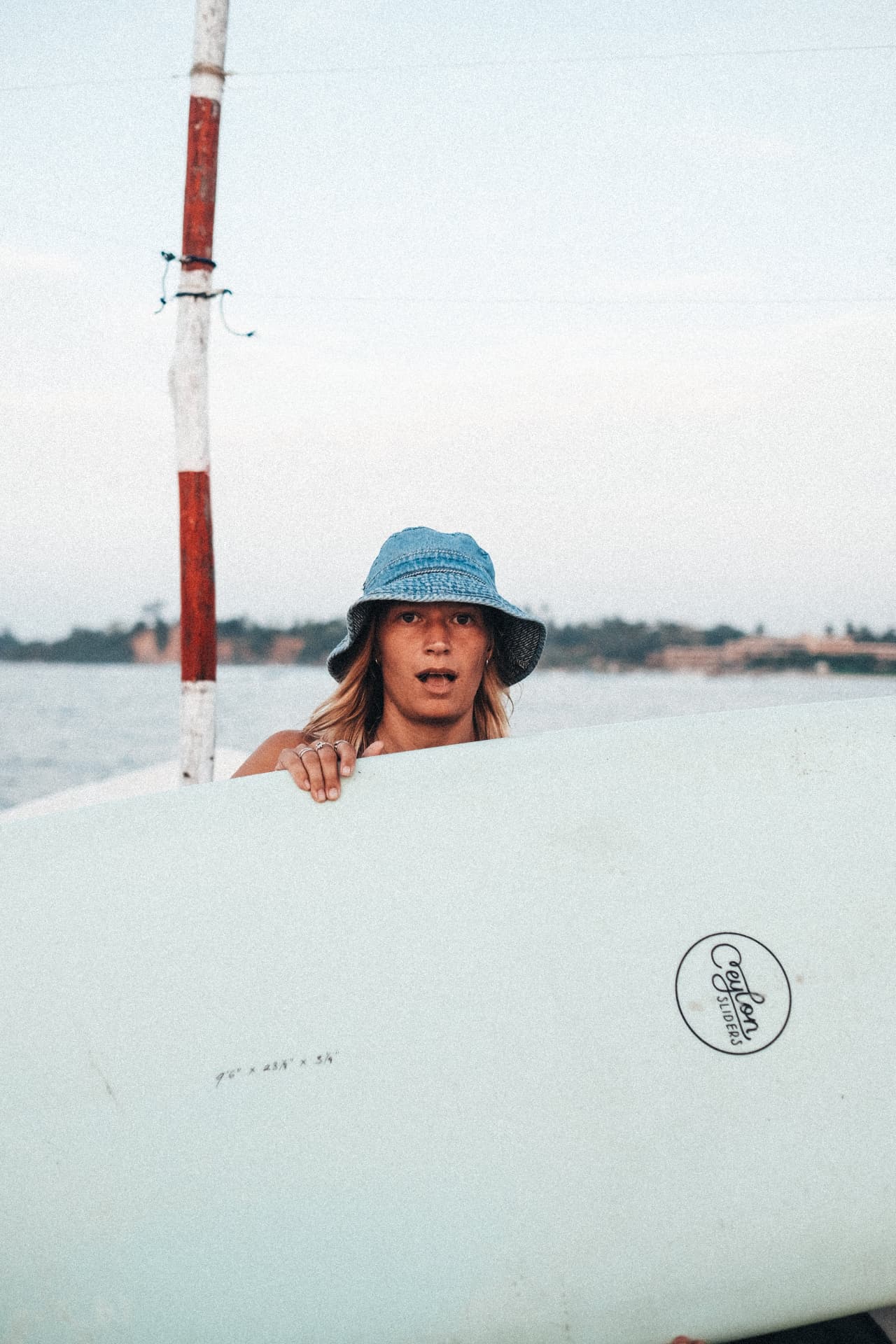 A surfer in a denim bucket hat peeking over a longboard on a boat