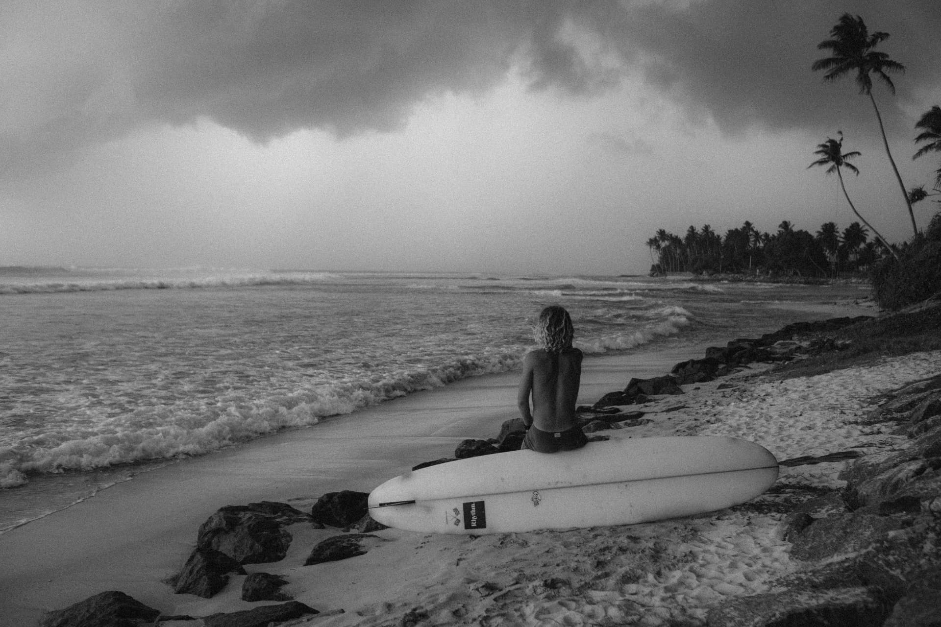 A surfer sitting on the sand with a longboard, watching the ocean