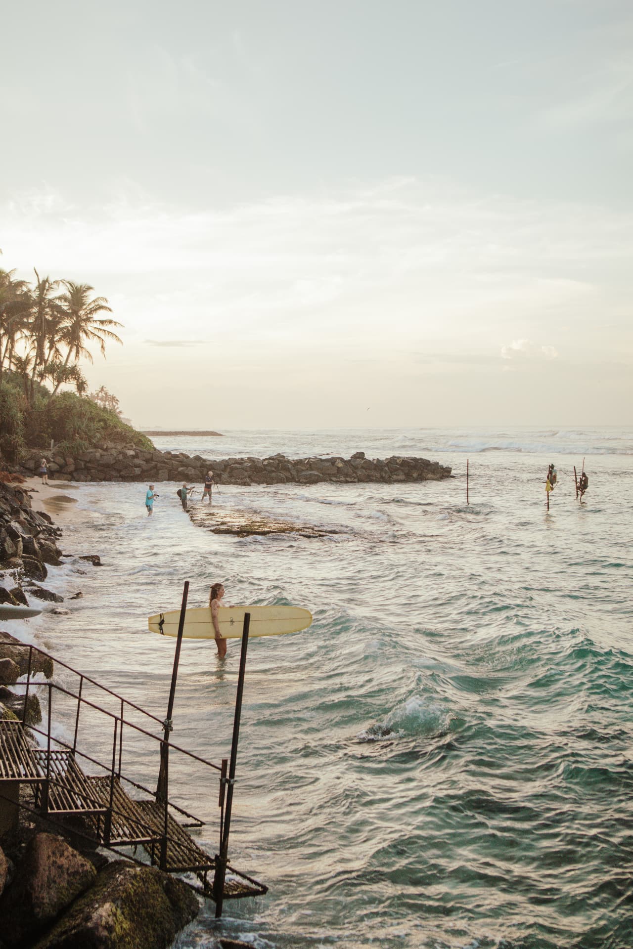 A surfer wading out past stilt fishermen at golden hour